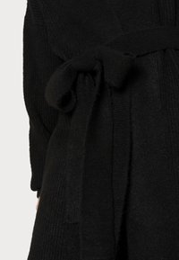 Czarny dziergany kardigan z paskiem w talii. Charakteryzuje się prążkowaną fakturą i miękkim wykończeniem. Pasek jest związany z przodu, zapewniając pewne zapięcie.