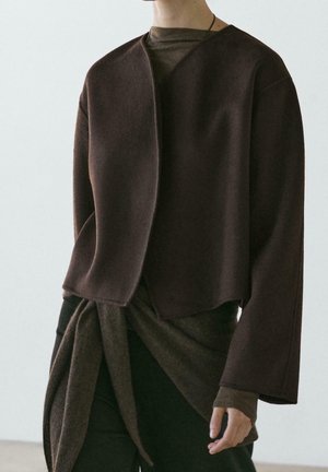 Persoon draagt een donkerbruine jas met open voorkant over een strakke, langarmige top en zwarte broek tegen een egale achtergrond.