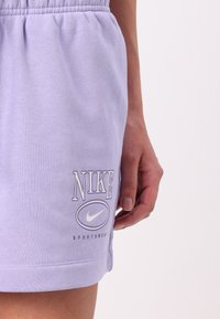 Shorts Nike lavande avec taille élastique, portant le logo blanc et le texte 'NIKE SPORTSWEAR', fabriqués en coton doux.