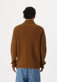Brauner gerippter Strickpullover mit hohem Kragen und langen Ärmeln. Der Stoff ist weich und dick und ergänzt locker sitzende blaue Jeansshorts.
