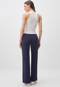 Donna in piedi di spalle, che indossa un top bianco senza maniche, pantaloni larghi blu navy e sneaker bianche su uno sfondo semplice.
