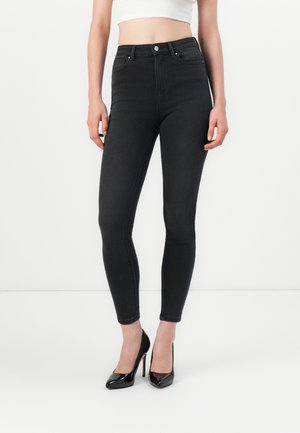 Zwarte high-waisted skinny jeans met een gladde textuur, voorzien van riemlussen, voorzakken en een nauwsluitend ontwerp, gecombineerd met zwarte hoge hakken.