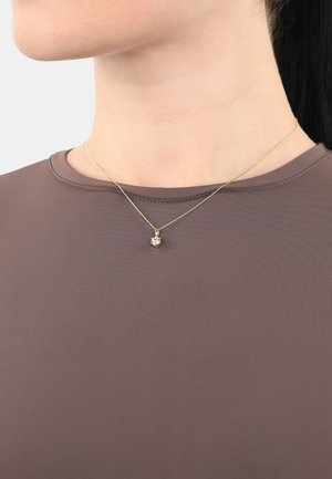 Gouden halsketting met een hanger met een heldere, veelkleurige steen. Draagt over een bruin, nauwsluitend shirt met een ronde hals.