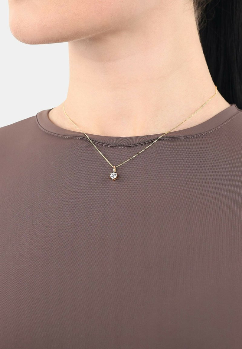 Collana d'oro con un pendente che presenta una pietra chiara e multifaccettata. Indossata sopra una camicia marrone aderente con scollatura rotonda.