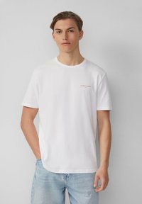 Jonge man met kort haar, gekleed in een wit T-shirt met kleine tekst op de borst en lichtblauwe versleten spijkerbroek, staande tegen een effen muur.