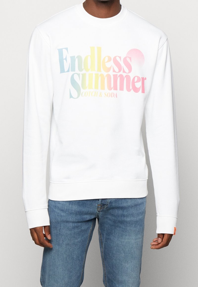 Homme portant un sweat-shirt blanc avec le texte pastel « Endless Summer Scotch & Soda » et un jean bleu, debout devant un fond uni.
