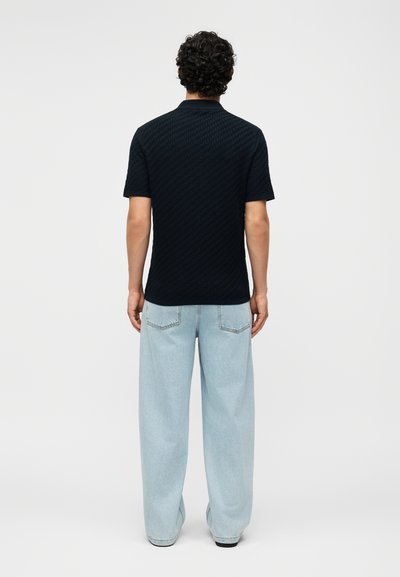 Άνδρας με σγουρά μαλλιά που φοράει μαύρο υφασμάτινο κοντομάνικο πουκάμισο και ανοιχτό μπλε τζιν με φαρδιά μπατζάκια, στέκεται με την πλάτη προς το απλό φόντο.