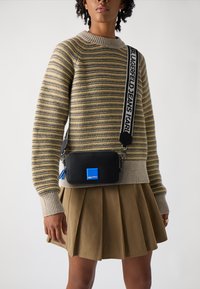 Geantă neagră tip crossbody cu accente albastre, textură de piele netedă, închidere cu fermoar și curea ajustabilă. Purtați-o cu un pulover cu dungi și o fustă plisată.