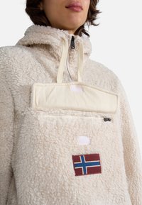 Kremowy polarowy pulower z wysokim kołnierzem, przednią naszywaną kieszenią i łatką z norweską flagą. Posiada zamek błyskawiczny oraz sznurek do regulacji.
