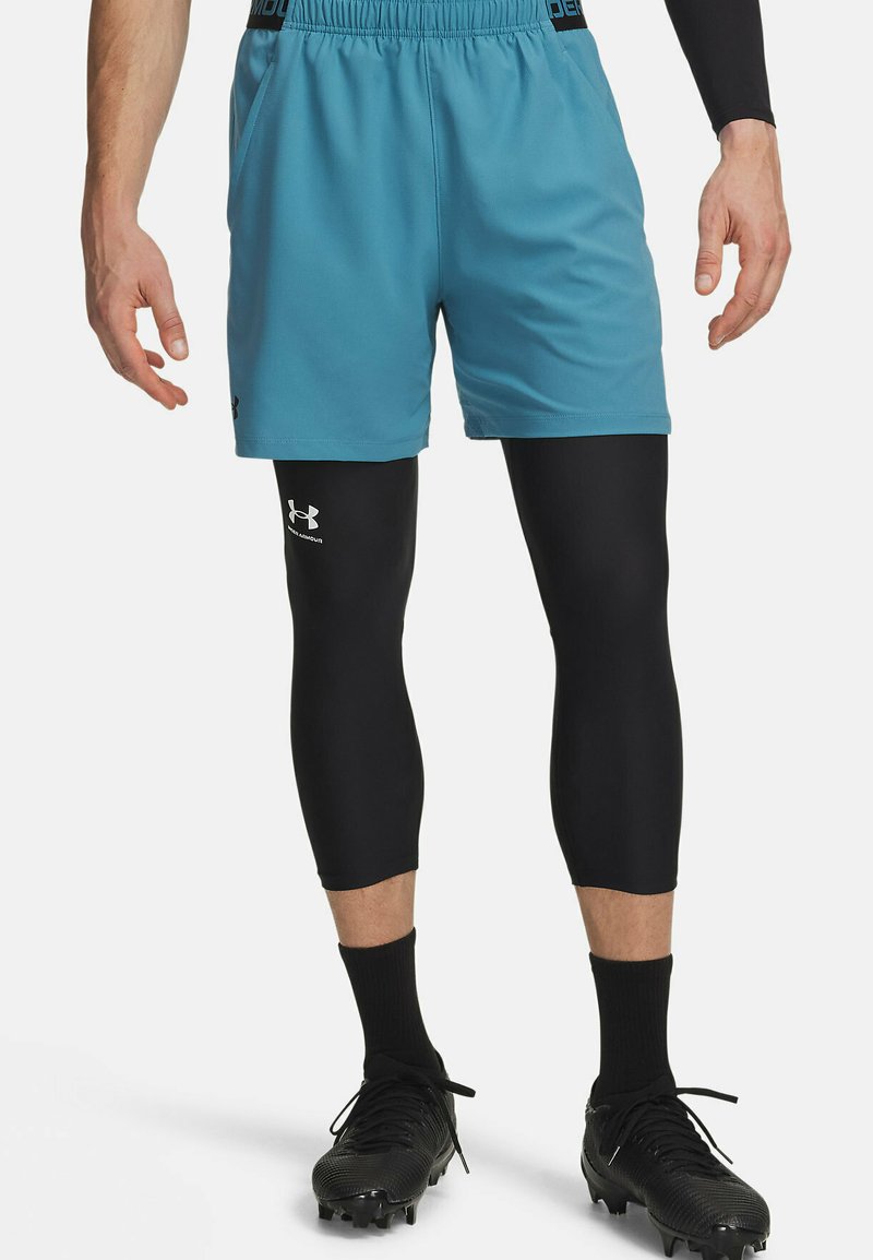 Türkisfarbene Sportshorts mit elastischem Bund, getragen über schwarzen Kompressionsleggings. Schwarze Stollenschuhe mit strukturiertem Grip.