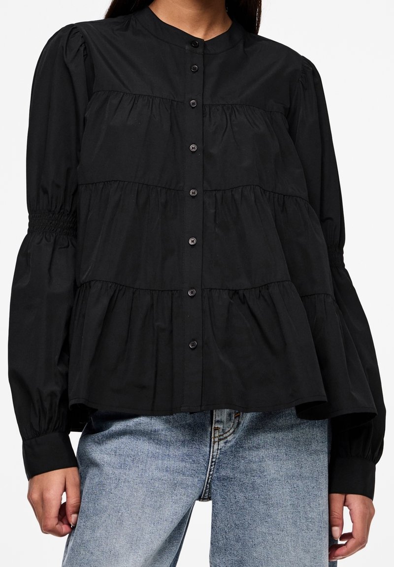 Blusa nera in cotone con una chiusura a bottoni frontale, volant arricciati e maniche a palloncino voluminose. Texture liscia con una silhouette strutturata.