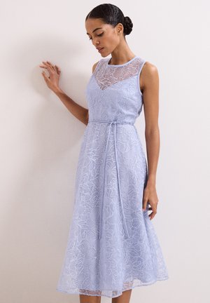 Frau trägt ein ärmelloses, helllila Spitzen-Midikleid mit Blumenmustern und einem dünnen Gürtel, steht vor einer schlichten hellen Wand.