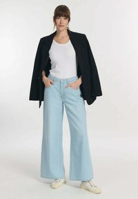 Lichtblauwe wijdekuit jeans, gecombineerd met een witte tanktop en een oversized zwarte blazer. Witte sneakers maken de outfit compleet. Soepele textuur, casual stijl.