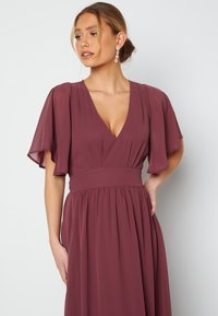 Mauve kjole med flagrende ærmer, dyb V-hals, tætsiddende talje og svajet nederdel. Glat stof med draperingsdetaljer, velegnet til formelle anledninger.