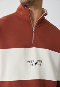 Sweatshirt cu fermoar de culoare ruginie, cu o dunga orizontală albă, având text brodat și emblema. Textură netedă, croială lejeră.