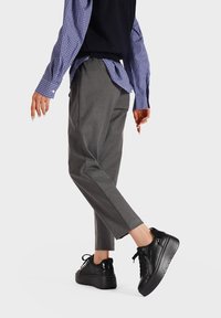 Sneakers platform neri con accenti lucidi, abbinati a pantaloni grigi su misura e una camicia a quadri blu indossata sotto un gilet nero.