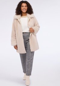 Beige kein keinäjärveen kanssa takki, jossa on huppu, napitettava sulku ja pehmeät hihansuut. Päällä valkoinen paita ja harmaaraidalliset ruutukankaat.