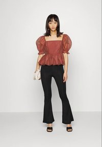 Mulher vestindo uma blusa peplum de manga bufante em cor ferrugem, calças pretas flare, sapatos pretos de dedo aberto, segurando uma clutch branca em fundo simples.