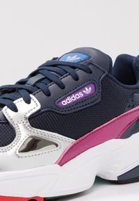 Mörkblå Adidas-sneakers med vit sula, lila logopatch, en rosa accentrand och metalliska silverdetaljer.