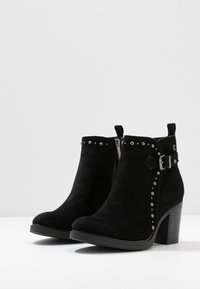 Botas de tobillo de ante negro con punta redonda, cremallera lateral, tachuelas decorativas alrededor del tobillo y un tacón grueso. El artículo cuenta con una hebilla lateral.