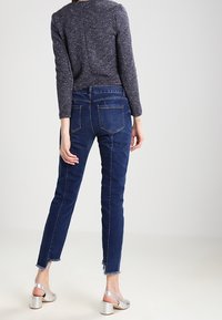 Donkerblauwe denim jeans met een slim fit en een rafelige zoom, gecombineerd met een gestructureerde navy top met lange mouwen. Zilveren sandalen met blokhak.