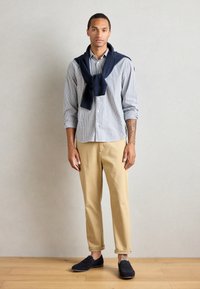 Cămașă cu dungi albă și albastră, pantaloni khaki tapered, pulover bleumarin aruncat pe umeri și loafers din suede închis. Fundal simplu.