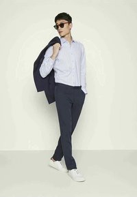 Un homme portant des lunettes de soleil, une chemise rayée blanche, un pantalon sombre, des baskets blanches, tenant une veste sombre sur son épaule, se tenant devant un fond clair.