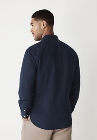 Marinblå långärmad skjorta med krage, knäppta manschetter och en slät tygstruktur, visad bakifrån.