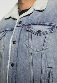 Hombre vistiendo una chaqueta de mezclilla azul clara con cuello de sherpa blanco y bolsillo en el pecho abotonado sobre una camisa negra.