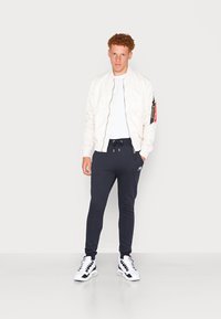 Letvægts, glansfuld hvid bomberjakke med lynlås; marineblå joggingbukser med snøre; sort og hvid sneakers; model står.