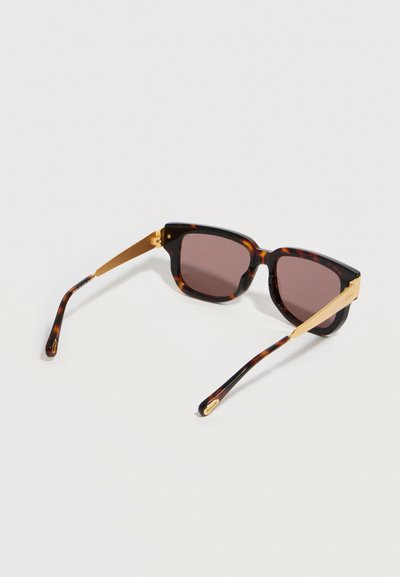 Γυαλιά ηλίου με καφέ τετράγωνα φακούς και χρυσούς μεταλλικούς βραχίονες σε σχέδιο χελώνας, εμφανιζόμενα από πίσω σε λευκό φόντο.