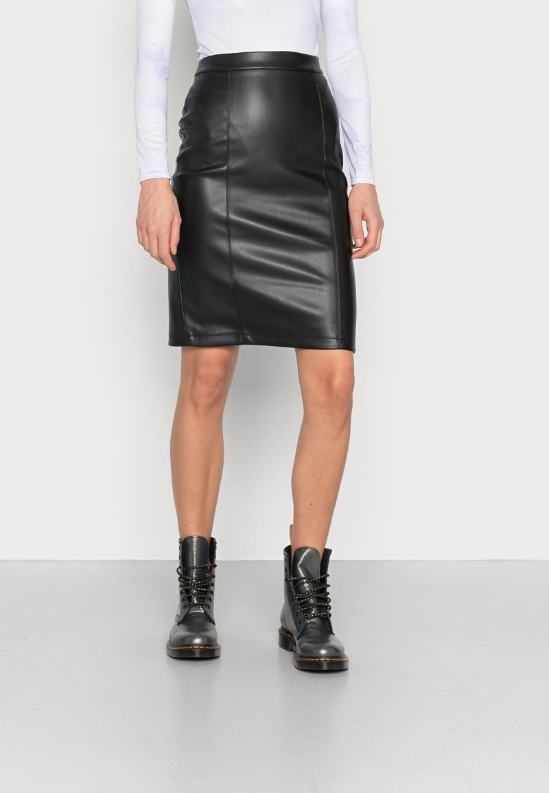 Femme portant une jupe crayon en cuir noire mi-longue, un haut blanc à manches longues et des bottines noires à lacets, debout sur un sol gris.