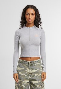 Top corto gris de manga larga con cuello alto y cremallera frontal, que presenta un logo rosa en el lado izquierdo del pecho; combinado con pantalones cargo de camuflaje.