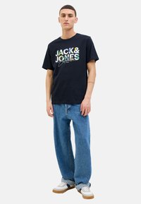 Navy Baumwoll-T-Shirt mit mehrfarbigem "JACK & JONES"-Logo, kombiniert mit hellblauen Jeans und weißen Sneakers mit braunen Sohlen.