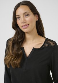 Haut noir à manches longues en tissu doux, avec un col en V et des accents en dentelle sur les épaules. Texture lisse avec une silhouette ajustée.