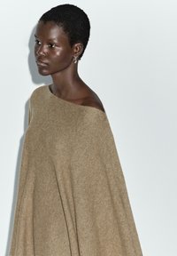 Pull beige hors-épaule avec une coupe ample et un tissu doux et texturé drapant élégamment, sur fond uni.