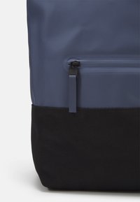 Sac à dos bleu marine en matériau lisse et imperméable, avec un fond en tissu noir, un compartiment principal et une petite poche zippée à l'avant.