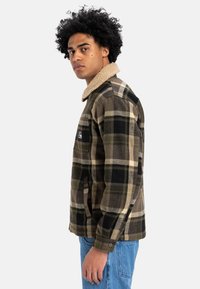 Rutet flanneljakke i oliven, sort og beige med en tan shearlingkrave. Har sidelommer og knapmanchetter. Holdbart materiale.