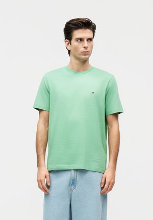 Mann trägt hellgrünes kurzärmeliges T-Shirt und hellblaue Jeans, steht vor schlichtem grauem Hintergrund und schaut nach rechts.