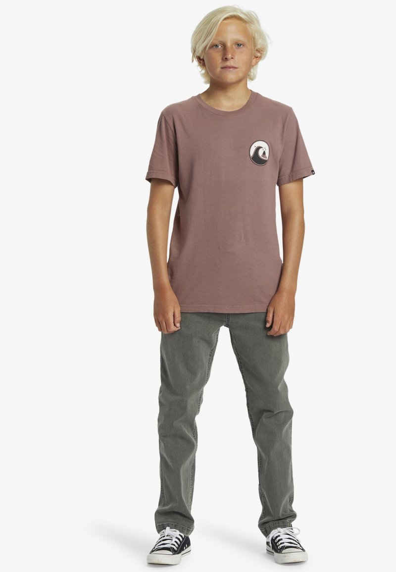 Camiseta de algodón marrón con mangas cortas y un logo de ola en blanco y negro en el pecho, combinada con pantalones gris y zapatillas negras.