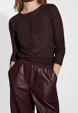 Femme portant un haut boutonné à manches longues marron foncé et un pantalon en cuir bordeaux taille haute avec les mains dans les poches.