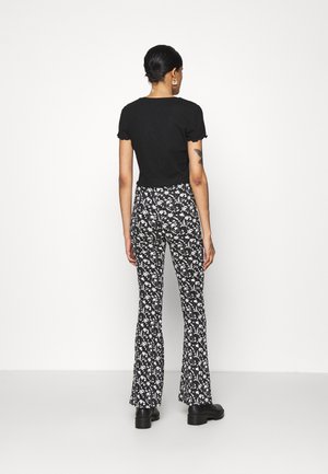 Donna con capelli neri raccolti in uno chignon, indossa una maglietta nera a maniche corte, pantaloni floreali in bianco e nero e scarpe nere con suola spessa, in piedi contro un muro bianco.