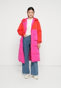 Parka lunga con parte superiore rossa e parte inferiore rosa, caratterizzata da bottoni a pressione e tasche a toppa. Indossata sopra una maglietta bianca e jeans.