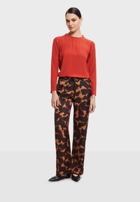 Crvena bluzica dugih rukava u kombinaciji sa crnim hlačama s apstraktnim uzorkom u narančastoj i smeđoj boji. Odjevna kombinacija uključuje pojas s vukučicom i elegantne cipele.