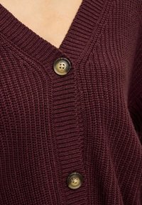 Gros plan sur une personne portant un cardigan tricoté bordeaux avec deux grands boutons marbrés le long de l'ouverture en V.