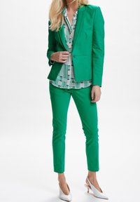 Zielona dopasowana marynarka i pasujące slim-fit spodnie, w połączeniu z wzorzystą bluzką. Białe szpilki w szpic dopełniają strój.