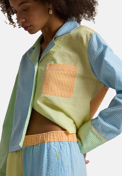 Femme portant une chemise rayée multicolore et un pantalon assorti avec une poche orange et une ceinture, debout dans une pose détendue devant un fond blanc.