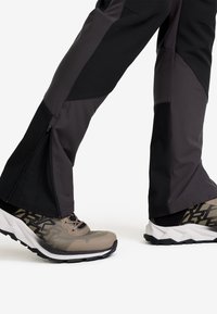 Svarta och grå hybridhängslen med zip-försedda instick, i kombination med ljusbruna sportskor med mesh-övre och mönstrade detaljer.