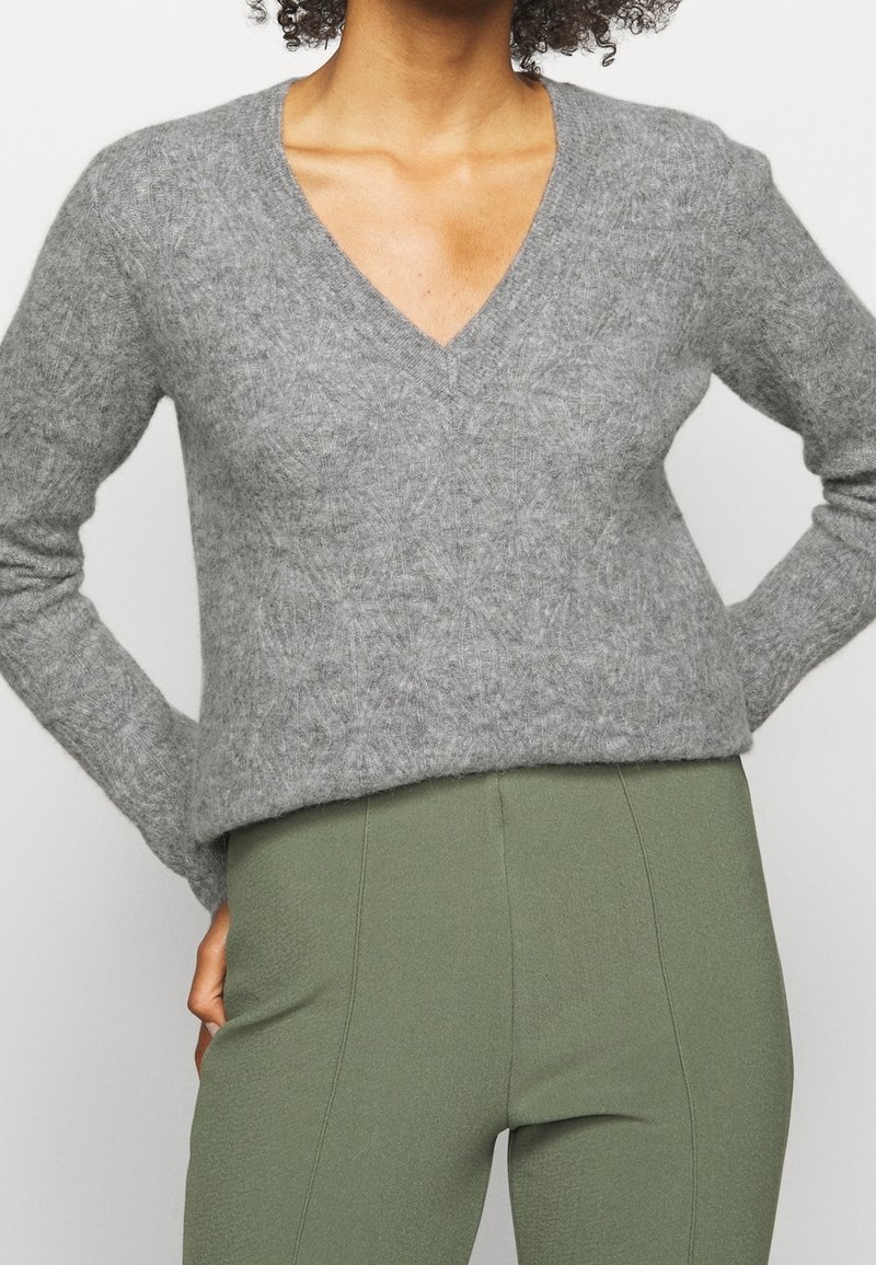 Personne portant un pull gris à col en V duveteux et un pantalon taille haute vert olive, les mains posées sur les hanches sur un fond neutre.