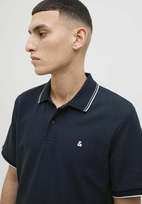 Tumes sinine polo-särk tekstuurse kangaga, valgete triipudega krae ja väike valge logo rindadel. Lühikesed varrukad, klassikaline lõige.